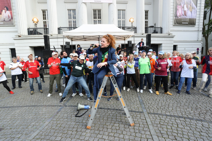 Vor dem Theater stehen sehr viele Menschen. Alle tragen Fußballtrikots. Vor ihnen steht eine Schauspielerin auf einer Leiter. Vor dem Theater stehen sehr viele Menschen. Alle tragen Fußballtrikots. Vor ihnen steht eine Schauspielerin auf einer Leiter.