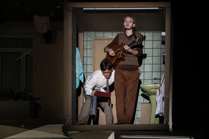 In einem sehr kleinen Badezimmer steht ein Musiker und spielt Gitarre. Ein Schauspieler steht gebückt neben ihm und versucht ihm etwas in die Hosentasche zu stecken. In einem sehr kleinen Badezimmer steht ein Musiker und spielt Gitarre. Ein Schauspieler steht gebückt neben ihm und versucht ihm etwas in die Hosentasche zu stecken.