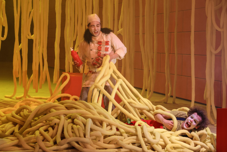Ein Sänger liegt auf dem Boden. Er ist wie ein Clown geschminkt und liegt inmitten von gelben Schläuchen. Die erinnern an riesige Spaghetti. Hinter ihm steht ein Sänger im Kostüm eines Kochs. Ein Sänger liegt auf dem Boden. Er ist wie ein Clown geschminkt und liegt inmitten von gelben Schläuchen. Die erinnern an riesige Spaghetti. Hinter ihm steht ein Sänger im Kostüm eines Kochs.