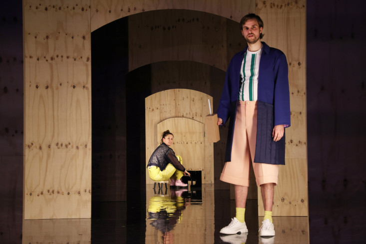 Eine Schauspielerin hockt vor dem winzigen Klavier. Vorn steht ein Schauspieler mit einem übergroßen Trinkpäckchen in der Hand. Eine Schauspielerin hockt vor dem winzigen Klavier. Vorn steht ein Schauspieler mit einem übergroßen Trinkpäckchen in der Hand.