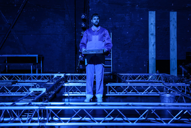 Ein Schauspieler hält einen Pizzakarton. Die Bühne ist in blaues Licht getaucht. Ein Schauspieler hält einen Pizzakarton. Die Bühne ist in blaues Licht getaucht.