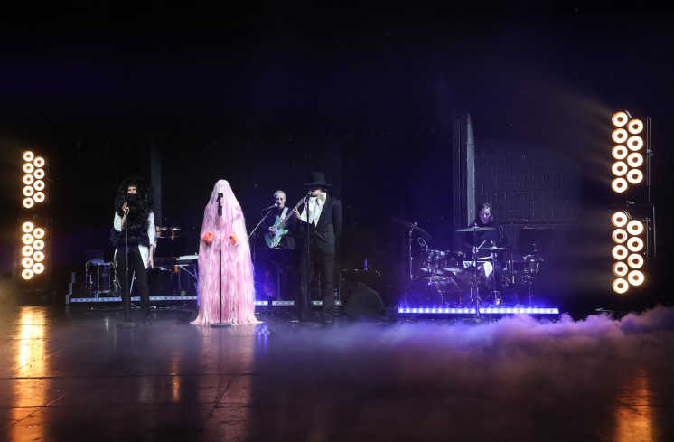 Eine Konzertsituation. Die Band sitzt auf der Bühne. Vor ihr stehen drei Personen an Mikrofonen. Eine trägt ein schwarzes Fellkostüm. In der Mitte steht jemand in einem pinken Ganzkörper-Fellkostüm. Eine Konzertsituation. Die Band sitzt auf der Bühne. Vor ihr stehen drei Personen an Mikrofonen. Eine trägt ein schwarzes Fellkostüm. In der Mitte steht jemand in einem pinken Ganzkörper-Fellkostüm.