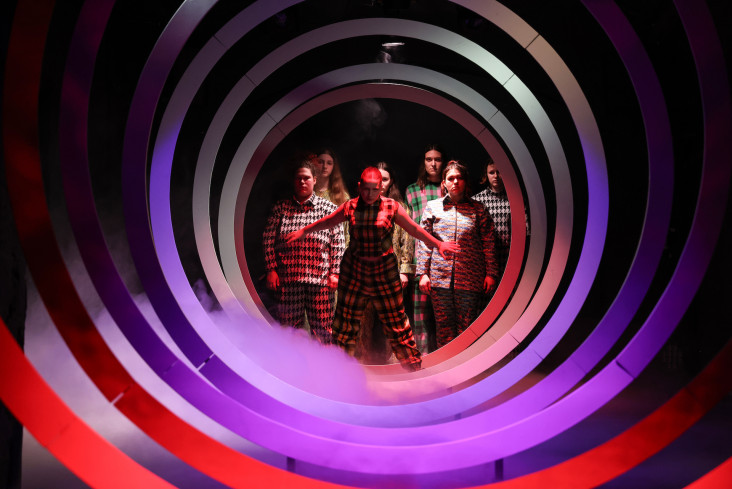 Das Bühnenbild sieht aus wie eine nach hinten zulaufende Spirale. Ganz hinten stehen sieben Personen. Die Ringe der Spirale sind in verschiedenen Farben angestrahlt.