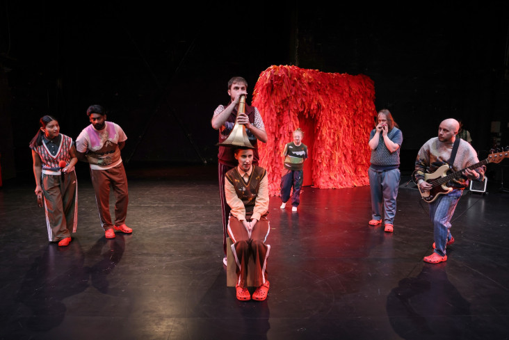 In der Mitte der Bühne sitzt eine Schauspielerin. Über ihr spricht eine Person in einen Trichter, der über ihrem Kopf schwebt. Um sie herum sind weitere fünf Personen. In der Mitte der Bühne sitzt eine Schauspielerin. Über ihr spricht eine Person in einen Trichter, der über ihrem Kopf schwebt. Um sie herum sind weitere fünf Personen.