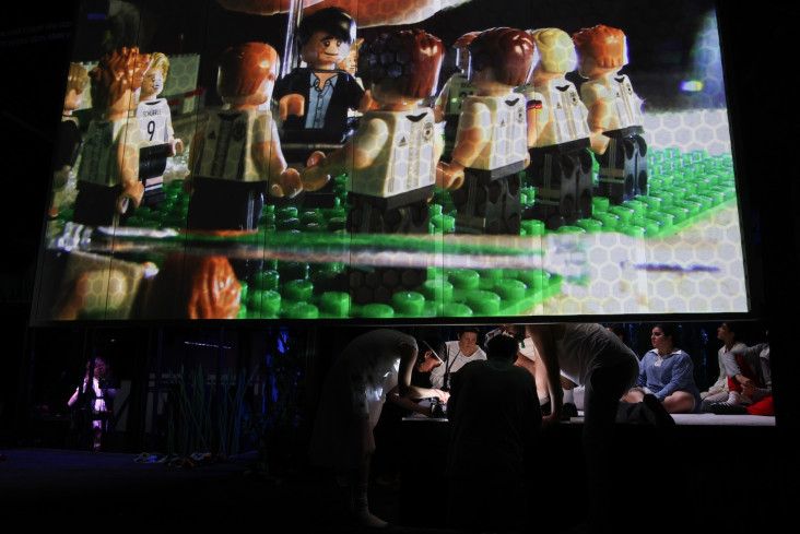 Auf einer großen Leinwand sind viele Legofiguren. Alle tragen Fußballtrikots. Hinter der Leinwand hocken mehrere Personen und spielen mit Legofiguren. Eine Person filmt. Auf einer großen Leinwand sind viele Legofiguren. Alle tragen Fußballtrikots. Hinter der Leinwand hocken mehrere Personen und spielen mit Legofiguren. Eine Person filmt.