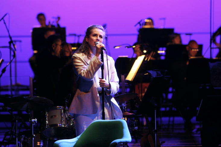 Nahaufnahme einer Sängerin, sie hält den Mikrofonständer mit beiden Händen fest und singt. Im Hintergrund spielt ein Orchester.