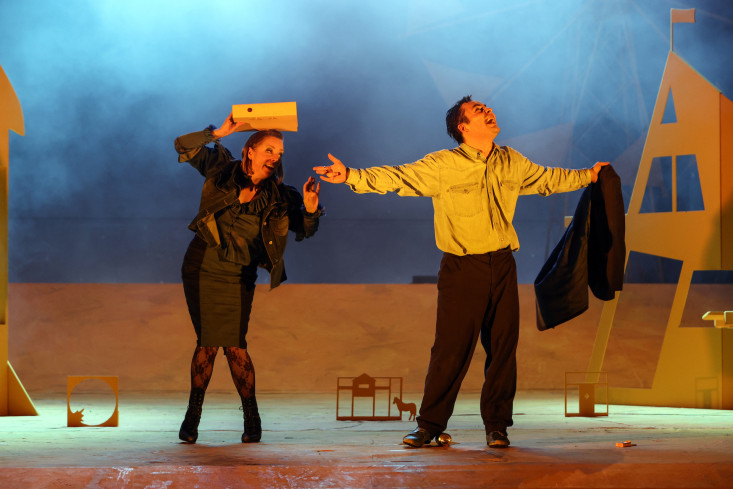 Eine Schauspielerin hält sich eine Aktenmappe über den Kopf. Sie streckt ihre Hand zu dem Schauspieler neben ihr aus. Der steht mit ausgebreiteten Armen da. Der Bühnenboden ist gelb, der Hintergrund blau beleuchtet. Eine Schauspielerin hält sich eine Aktenmappe über den Kopf. Sie streckt ihre Hand zu dem Schauspieler neben ihr aus. Der steht mit ausgebreiteten Armen da. Der Bühnenboden ist gelb, der Hintergrund blau beleuchtet.