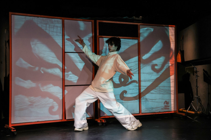 Eine hell gekleidete Person tanzt. Hinter dem Tänzer sind Projektionen auf einer Wand. Eine hell gekleidete Person tanzt. Hinter dem Tänzer sind Projektionen auf einer Wand.