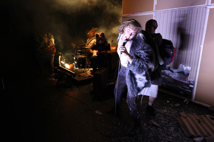 Eine Frau in einem Fellmantel singt. Hinter ihr spielt eine Band. Alle drei Musiker tragen große Tiermasken. Eine Frau in einem Fellmantel singt. Hinter ihr spielt eine Band. Alle drei Musiker tragen große Tiermasken.