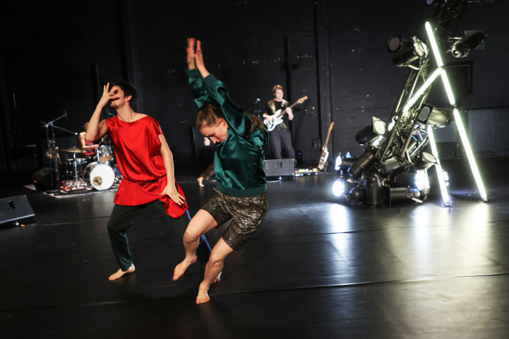 Vorn tanzen zwei Personen. Der Tänzer trägt ein rotes Oberteil, die Tänzerin ein grünes. Hinten sind Musiker zu sehen. Vorn tanzen zwei Personen. Der Tänzer trägt ein rotes Oberteil, die Tänzerin ein grünes. Hinten sind Musiker zu sehen.