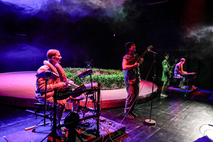 Das Schauspielensemble steht vorn an der Bühne. Eine Person sitzt an einem Schlagzeug, eine an einem Keyboard. Die anderen beiden spielen Bass oder E-Gitarre. Das Schauspielensemble steht vorn an der Bühne. Eine Person sitzt an einem Schlagzeug, eine an einem Keyboard. Die anderen beiden spielen Bass oder E-Gitarre.