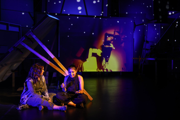 Zwei Schauspielerinnen sitzen im Schneidersitz auf dem Boden. Auf die Wand hinter ihnen sind Farben und Formen projiziert. Zwei Schauspielerinnen sitzen im Schneidersitz auf dem Boden. Auf die Wand hinter ihnen sind Farben und Formen projiziert.