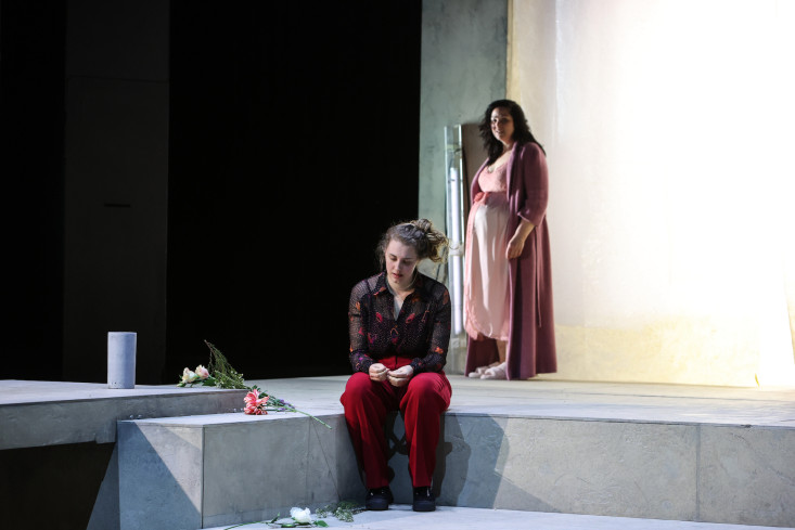 Zwei Schauspielerinnen sind auf der Bühne. Die eine sitzt vorn auf dem Boden. Die zweite steht weiter hinten. Sie trägt rosafarbene Nachtwäsche und hat einen Babybauch. Zwei Schauspielerinnen sind auf der Bühne. Die eine sitzt vorn auf dem Boden. Die zweite steht weiter hinten. Sie trägt rosafarbene Nachtwäsche und hat einen Babybauch.