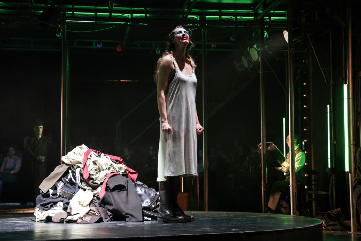 Eine Schauspielerin steht neben einem Kleiderberg. Sie trägt ein weißes, einfaches Kleid und schaut nach oben. Eine Schauspielerin steht neben einem Kleiderberg. Sie trägt ein weißes, einfaches Kleid und schaut nach oben.
