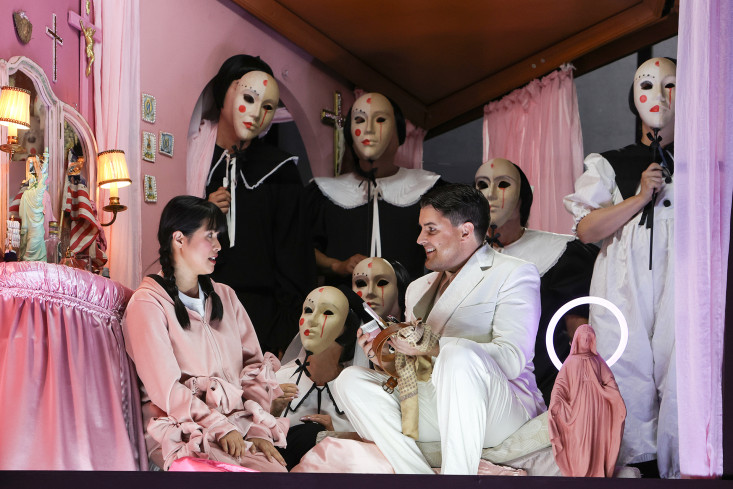 In einem komplett rosa ausstaffierten Raum sitzen sich eine Sängerin und ein Sänger gegenüber. Um sie herum stehen sehr viele Personen mit Masken. In einem komplett rosa ausstaffierten Raum sitzen sich eine Sängerin und ein Sänger gegenüber. Um sie herum stehen sehr viele Personen mit Masken.