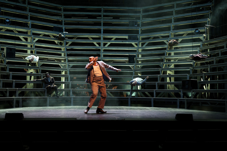 Ein Mann steht auf der Bühne. Das Bühnenbild ist eine Art Arena mit steil aufsteigenden Streben.  Ein Mann steht auf der Bühne. Das Bühnenbild ist eine Art Arena mit steil aufsteigenden Streben.