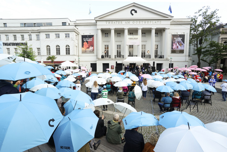 Sehr viele Menschen sitzen mit aufgespannten Regenschirmen auf dem Goetheplatz. Es regnet nicht. Im Hintergrund ist das Theater. Sehr viele Menschen sitzen mit aufgespannten Regenschirmen auf dem Goetheplatz. Es regnet nicht. Im Hintergrund ist das Theater.