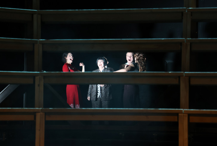 Hinter den Streben des Bühnenbilds stehen mehrere Personen. Sie lachen mit weit geöffneten Mündern. Hinter den Streben des Bühnenbilds stehen mehrere Personen. Sie lachen mit weit geöffneten Mündern.