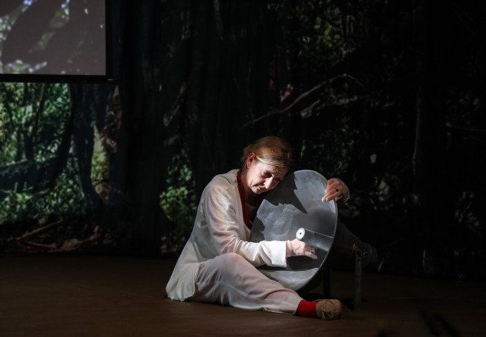Eine Schauspielerin sitzt auf dem Boden. Sie hält einen großen Lautsprecher im Arm. Die Bühne ist dunkel. Eine Schauspielerin sitzt auf dem Boden. Sie hält einen großen Lautsprecher im Arm. Die Bühne ist dunkel.