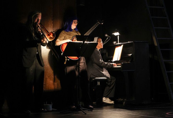 Die Band spielt Musik. Um sie herum ist es dunkel. Eine Person sitzt am Klavier, eine andere spielt Bass, einer Posaune.