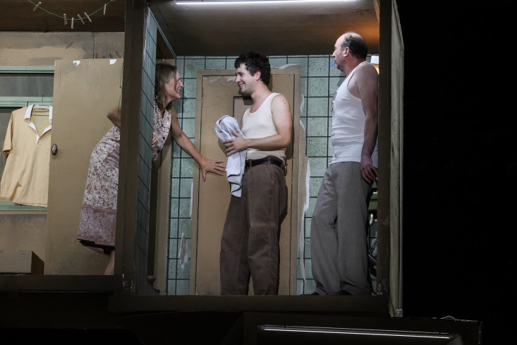 In einem sehr kleinen Badezimmer stehen zwei Schauspieler. Eine Schauspielerin schaut durch die Tür zu ihnen herein. In einem sehr kleinen Badezimmer stehen zwei Schauspieler. Eine Schauspielerin schaut durch die Tür zu ihnen herein.