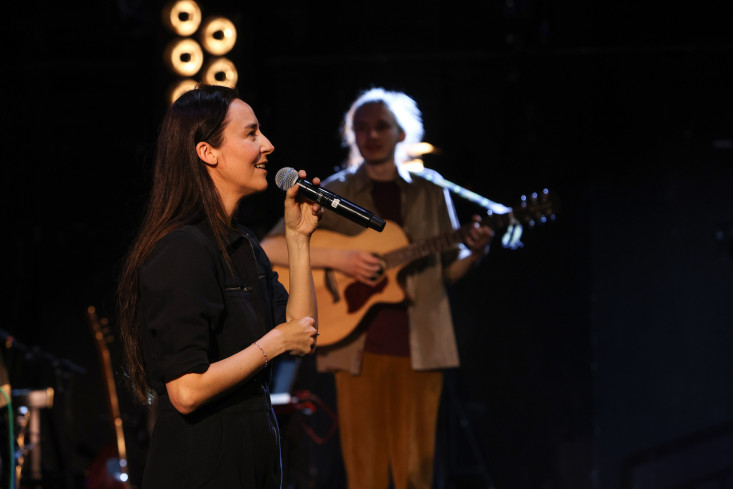 Eine Frau singt. Sie hält ein Mikrofon in der Hand. Hinten steht ein Musiker und spielt Gitarre.