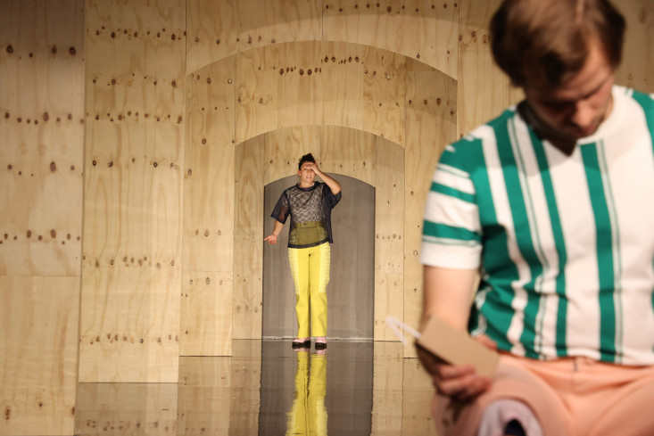 Eine Schauspielerin steht hinten auf der Bühne und hält die Hand über die Augen. Vorn sitzt ein Schauspieler mit einem Trinkpäckchen in der Hand.