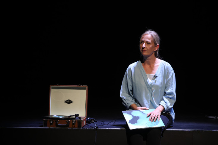 Eine Schauspielerin sitzt auf der Bühnenkante mit einer Schallplatte in der Hand. Neben ihr steht ein alter Plattenspieler. Eine Schauspielerin sitzt auf der Bühnenkante mit einer Schallplatte in der Hand. Neben ihr steht ein alter Plattenspieler.
