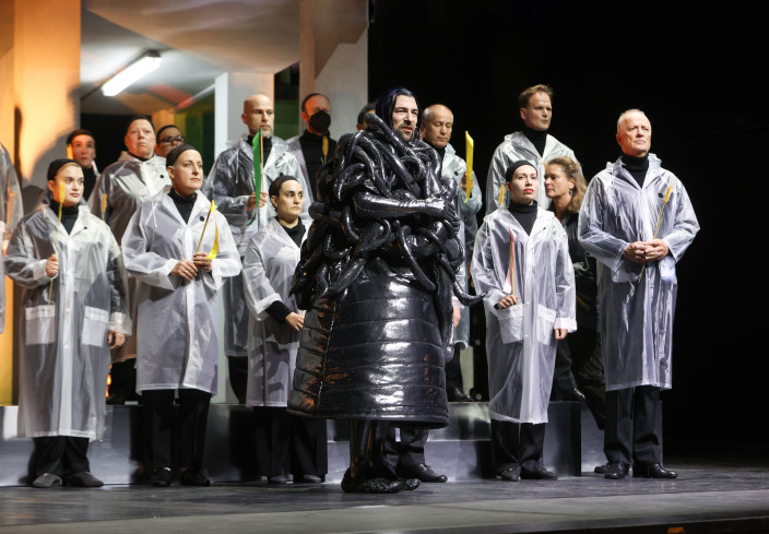 Vorn steht ein Schauspieler in einem schwarzen Kostüm. Sein Oberkörper ist von Schläuchen umwunden. Hinter ihm steht der Chor. Vorn steht ein Schauspieler in einem schwarzen Kostüm. Sein Oberkörper ist von Schläuchen umwunden. Hinter ihm steht der Chor.