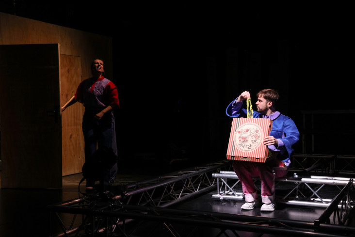 Ein Schauspieler sitzt und hält einen Pizzakarton auf den Knien. Der ist geöffnet. Der Schauspieler holt gelbe Luftschlangen daraus. Neben ihm steht eine Schauspielerin. Sie schaut nach oben zur Decke. Ein Schauspieler sitzt und hält einen Pizzakarton auf den Knien. Der ist geöffnet. Der Schauspieler holt gelbe Luftschlangen daraus. Neben ihm steht eine Schauspielerin. Sie schaut nach oben zur Decke.