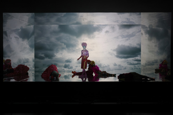 Vier Personen liegen auf der Bühne. In der Mitte steht eine Schauspielerin. Auf die Bühne sind Wolken projeziert. Vier Personen liegen auf der Bühne. In der Mitte steht eine Schauspielerin. Auf die Bühne sind Wolken projeziert.