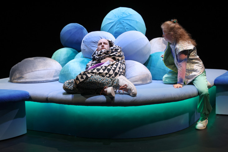 Ein Schauspieler liegt auf vielen Kissen. Er ist zur Seite gedreht. Hinter ihm sitzt eine Schauspielerin. Sie sieht ihn an. Ein Schauspieler liegt auf vielen Kissen. Er ist zur Seite gedreht. Hinter ihm sitzt eine Schauspielerin. Sie sieht ihn an.
