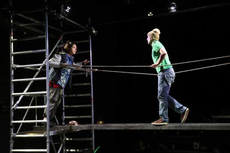 Eine Schauspielerin lehnt oben im Gerüst, sie diskutiert mit dem Schauspieler, der über eine Brücke auf sie zukommt. Eine Schauspielerin lehnt oben im Gerüst, sie diskutiert mit dem Schauspieler, der über eine Brücke auf sie zukommt.