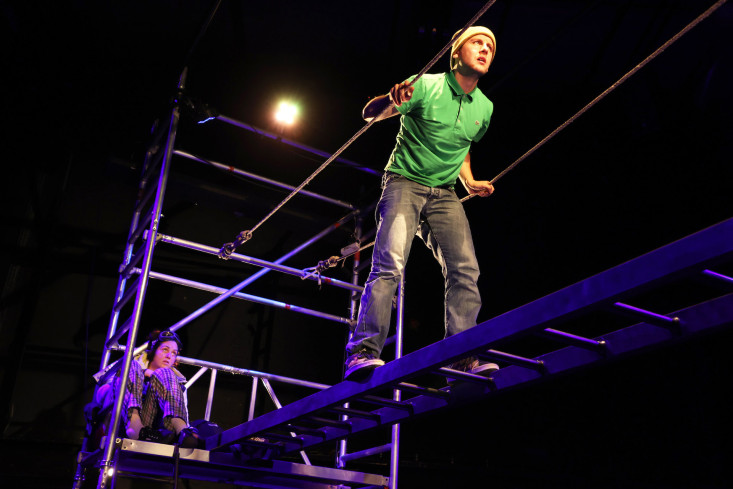 Ein Schauspieler läuft oben auf dem Gerüst über eine Brücke. An deren einem Ende sitzt mit angezogenen Beinen eine Schauspielerin und schaut ihm zu. Ein Schauspieler läuft oben auf dem Gerüst über eine Brücke. An deren einem Ende sitzt mit angezogenen Beinen eine Schauspielerin und schaut ihm zu.