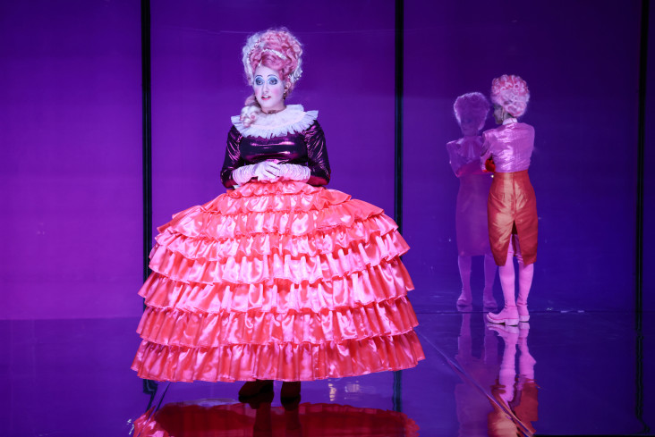 Eine Schauspielerin steht vorn. Sie trägt einen pinken Reifrock. Hinten steht eine andere Schauspielerin. Sie kehrt dem Publikum den Rücken zu. Eine Schauspielerin steht vorn. Sie trägt einen pinken Reifrock. Hinten steht eine andere Schauspielerin. Sie kehrt dem Publikum den Rücken zu.