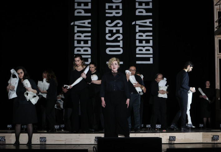 In der Mitte der Bühne steht eine schwarz gekleidete Sängerin. Hinter ihr sind mehrere schwarz gekleidete Personen. Alle halten weiße Stücke von Schaufensterpuppen in den Händen.