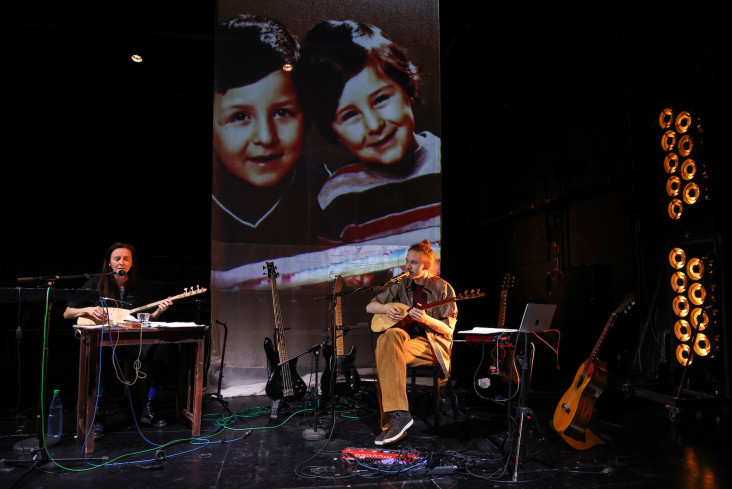 Auf einer großen Leinwand ist ein Bild von zwei Kindern zu sehen. Davor sitzen eine Frau und ein Mann. Beide spielen ein Saiteninstrument. Neben ihnen stehen viele andere Instrumente.