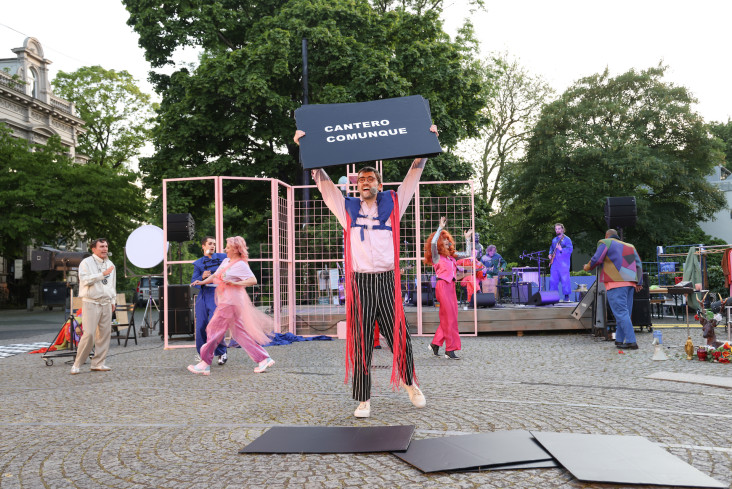 Sechs Personen sind auf dem Goetheplatz. Vorn steht ein Mann mit einem hocherhobenen Schild. Auf dem steht Cantero Comunque. Alle sind farbenfroh angezogen und sehr beschäftigt. Hinten spielt die Band.