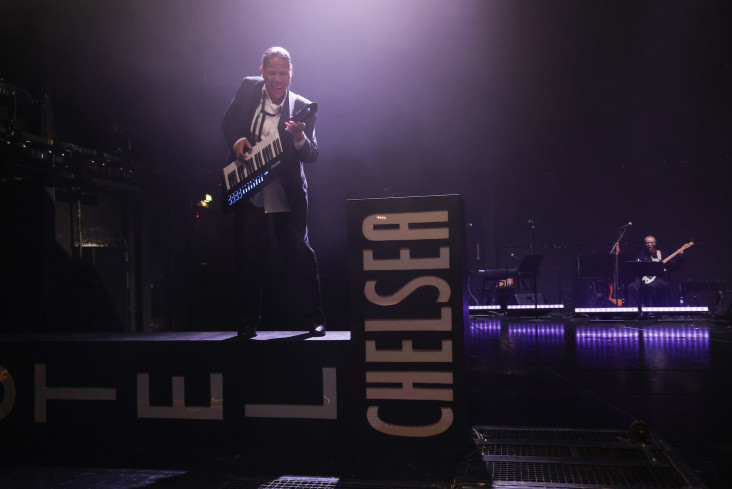 Die Bühne ist wie bei einem Konzert beleuchtet. Eine Musikerin steht im Scheinwerferlicht. Sie steht auf einem umgestürzten Schild, auf dem Hotel Chelsea zu lesen ist. Die Bühne ist wie bei einem Konzert beleuchtet. Eine Musikerin steht im Scheinwerferlicht. Sie steht auf einem umgestürzten Schild, auf dem Hotel Chelsea zu lesen ist.