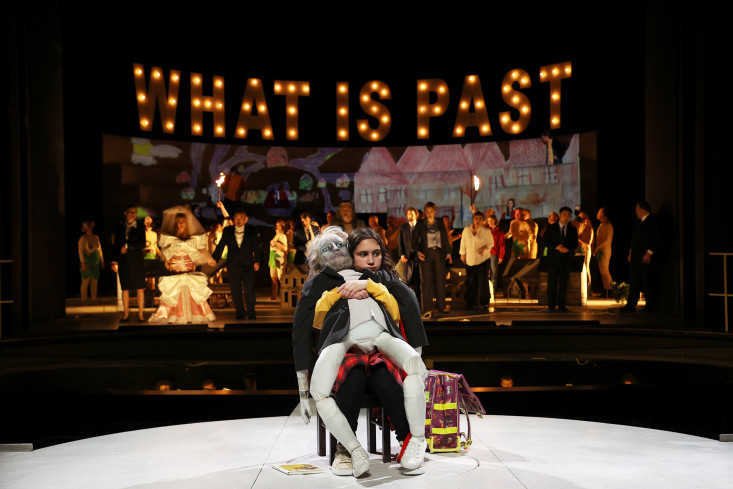 Ein Kind mit einer großen Falstaff-Puppe sitzt auf dem Steg im Zuschauerraum. Auf der Bühne hinter ihm ist eine große Gesellschaft. Ein Kind mit einer großen Falstaff-Puppe sitzt auf dem Steg im Zuschauerraum. Auf der Bühne hinter ihm ist eine große Gesellschaft.