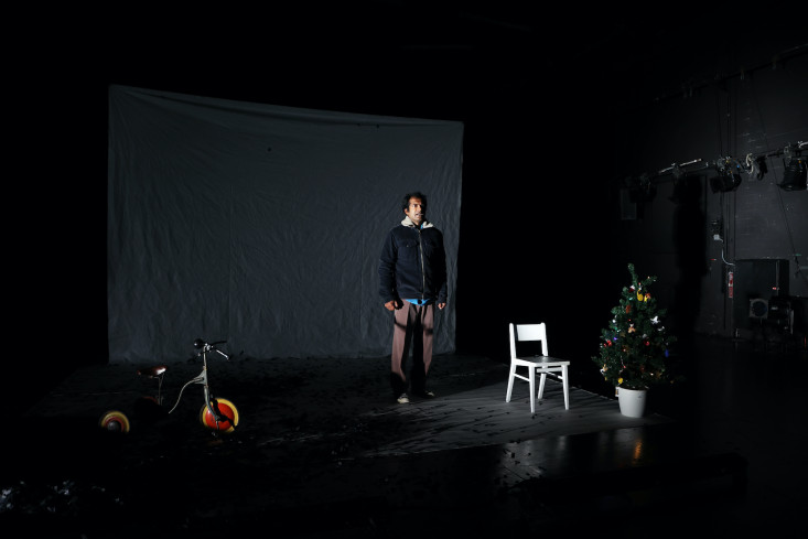 Die Bühne ist dunkel und karg. Links steht ein Dreirad, rechts ein weißer Stuhl und ein kleiner Weihnachtsbaum. In der Mitte steht mit hängenden Armen und in Winterjacke der Schauspieler. Die Bühne ist dunkel und karg. Links steht ein Dreirad, rechts ein weißer Stuhl und ein kleiner Weihnachtsbaum. In der Mitte steht mit hängenden Armen und in Winterjacke der Schauspieler.