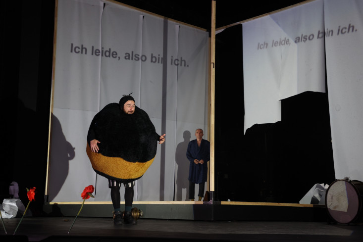 Ein Schauspieler in einem plüschigen Bienenkostüm steht auf der Bühne. Auf eine Papierbahn hinter ihm ist ein Satz projeziert: Ich leide, also bin ich. Ein Schauspieler in einem plüschigen Bienenkostüm steht auf der Bühne. Auf eine Papierbahn hinter ihm ist ein Satz projeziert: Ich leide, also bin ich.