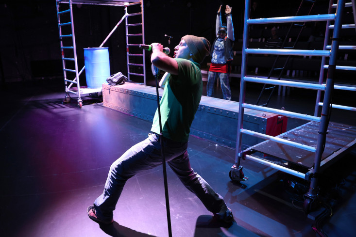 © Jörg Landsberg | Ein Schauspieler steht breitbeinig und singt in ein Mikrofon. Hinten steht eine Schauspielerin und hüpft. Ein Schauspieler steht breitbeinig und singt in ein Mikrofon. Hinten steht eine Schauspielerin und hüpft.
