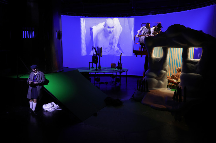 Die ganze Bühne ist auf dem Foto. Rechts ein kleines Häuschen, in dem ein Schauspieler sitzt. Sein Gesicht wird per Video auf die große Wand hinten projeziert. Vorn steht eine Schauspielerin. Die ganze Bühne ist auf dem Foto. Rechts ein kleines Häuschen, in dem ein Schauspieler sitzt. Sein Gesicht wird per Video auf die große Wand hinten projeziert. Vorn steht eine Schauspielerin.