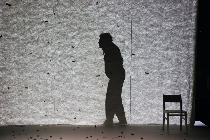 Der Schauspieler ist nur als gebeugter Schatten hinter der Leinwand zu sehen. Schwarze Papierschnipsel wehen durch die Luft und wirken in dem Schattenspiel wie Schneeflocken. Der Schauspieler ist nur als gebeugter Schatten hinter der Leinwand zu sehen. Schwarze Papierschnipsel wehen durch die Luft und wirken in dem Schattenspiel wie Schneeflocken.