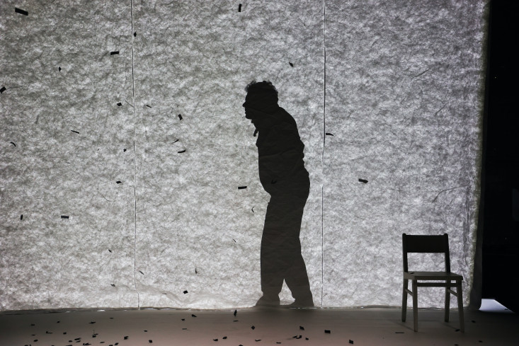 Der Schauspieler ist nur als gebeugter Schatten hinter der Leinwand zu sehen. Schwarze Papierschnipsel wehen durch die Luft und wirken in dem Schattenspiel wie Schneeflocken.