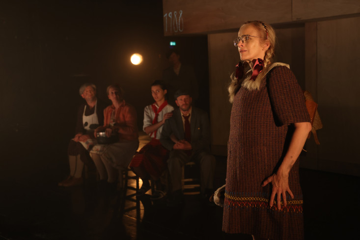 Vorn steht eine Schauspielerin mit geflochtenen Zöpfen und einem Kleid. Hinten sitzen vier Personen und betrachten sie. Vorn steht eine Schauspielerin mit geflochtenen Zöpfen und einem Kleid. Hinten sitzen vier Personen und betrachten sie.
