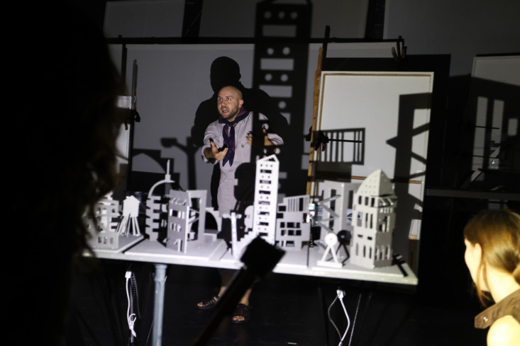 Auf einem Tisch ist eine kleine Stadt aufgebaut. Alles ist weiß. Hinter dem Tisch steht ein Schauspieler. Auf einem Tisch ist eine kleine Stadt aufgebaut. Alles ist weiß. Hinter dem Tisch steht ein Schauspieler.
