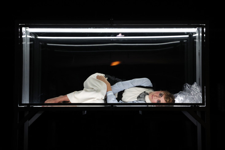 Eine Frau liegt in einem Glaskasten. Die Bühne hinter ihr ist schwarz. Eine Frau liegt in einem Glaskasten. Die Bühne hinter ihr ist schwarz.