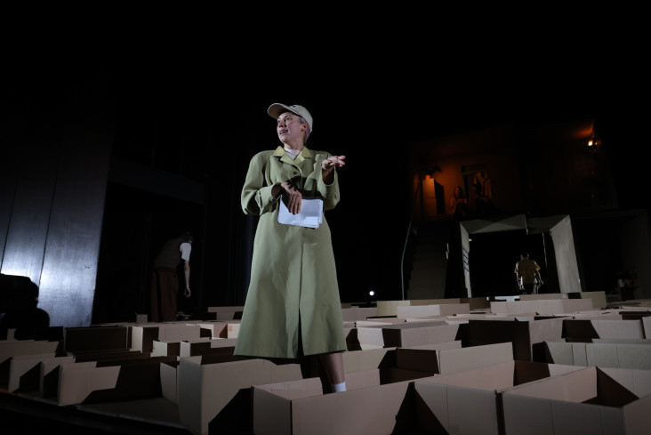 Eine Schauspielerin steht mitten zwischen Umzugskartons. Sie trägt einen Mantel und ein Basecap. In der Hand hält sie ein Blatt Papier. Eine Schauspielerin steht mitten zwischen Umzugskartons. Sie trägt einen Mantel und ein Basecap. In der Hand hält sie ein Blatt Papier.